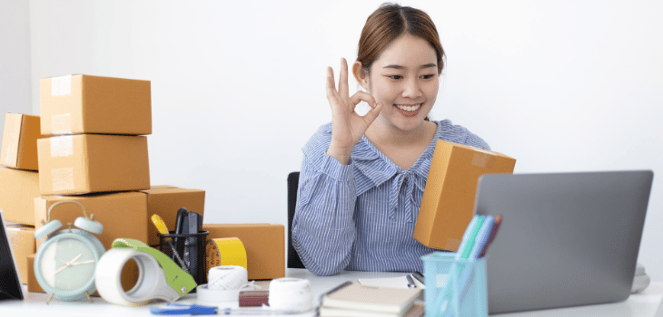 Imagem mostra mulher branca feliz na frente do notebook, com uma caixa de encomenda em uma das mãos e atrás também há caixas.