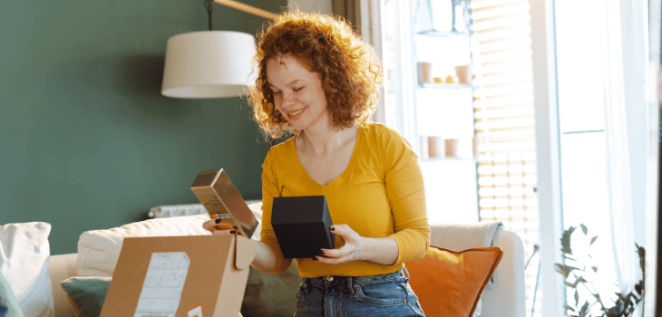 Mulher ruiva, com blusa laranja, com produtos nas mãos, sorrindo e uma caixa de papelão aberta em sua frente.