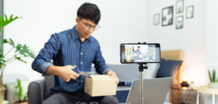Homem sentado em um sofá, usando camisa azul, abrindo uma caixa de papelão pequena com uma tesoura e um celular no tripé filmando.
